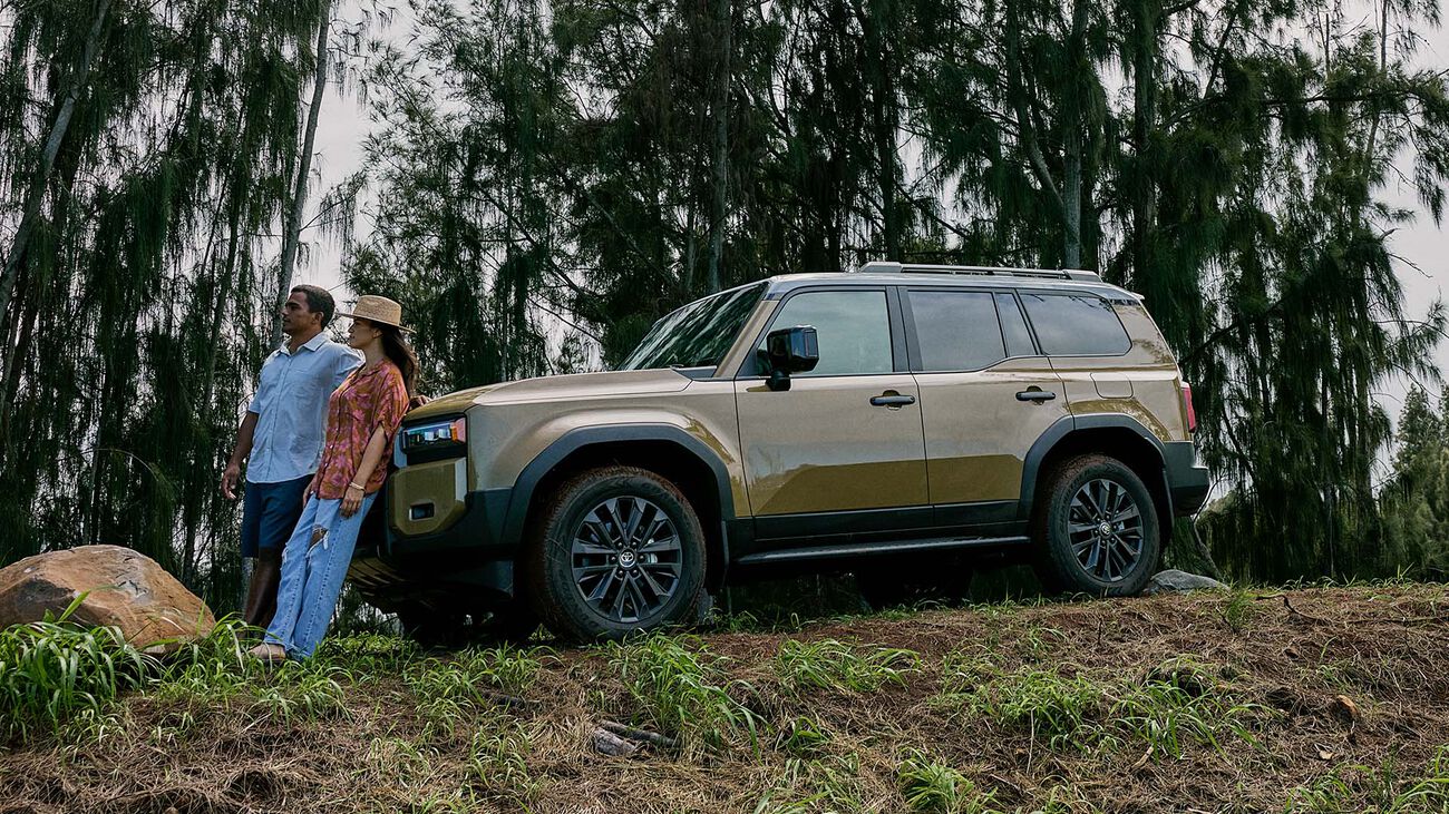 Toyota Land Cruiser in Trail Dust parked up on a hill with a young couple leaning against the front of the vehicle looking into the distance