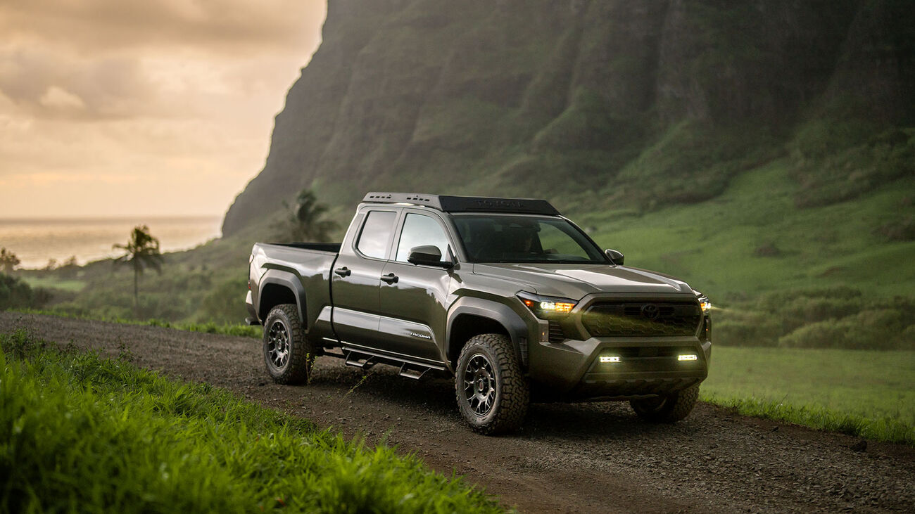 The 2026 Toyota Tacoma driving along a dirt path with luscious green mountains surrounding it.