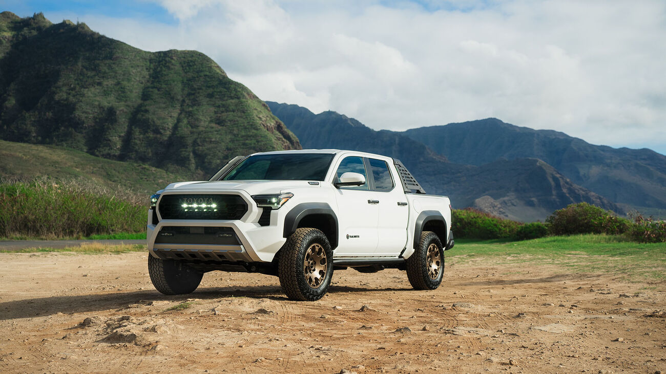 2026 Toyota Tacoma i-FORCE MAX driving on rugged terrain with green Hawaii mountains in the background.
