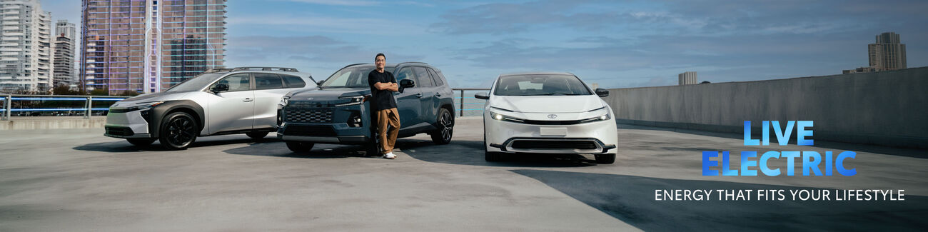 Young professional posed in front of the Toyota bZ Woodland, Prius Plug-in Hybrid, and RAV4 on the top of a parking structure.