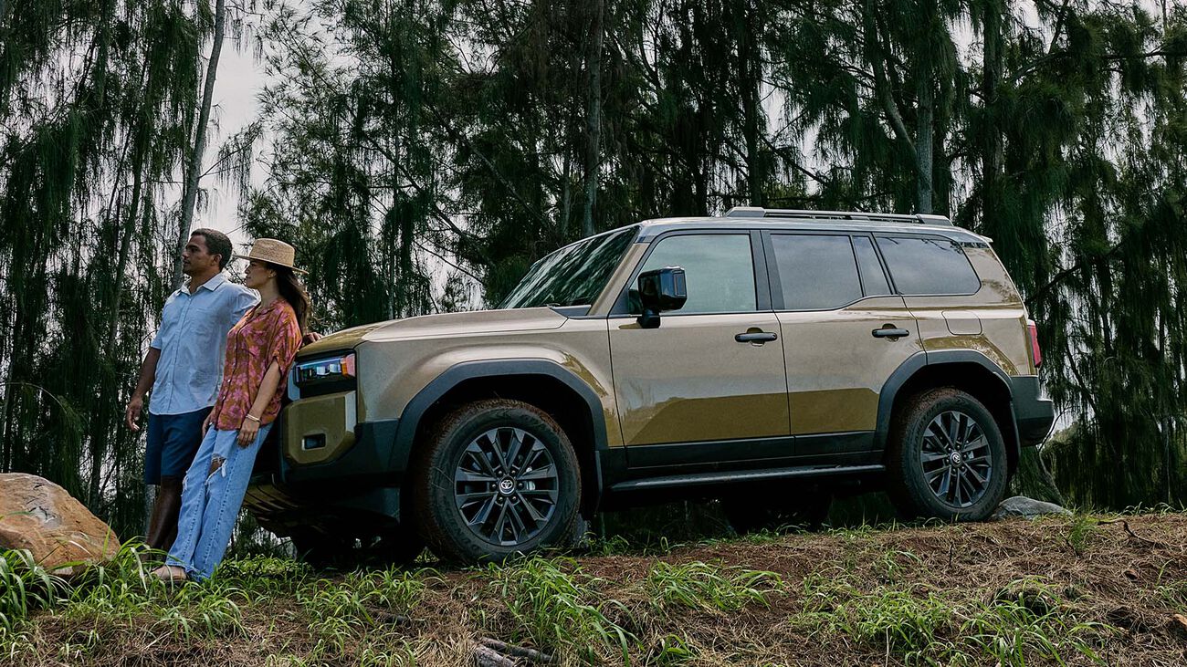 2026 Toyota Land Cruiser parked along a forest ridge with a  young couple leaning against the front of the vehicle.